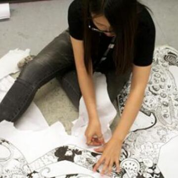 A student sitting on the floor drawing patterns on a large, organically cut out piece of paper.