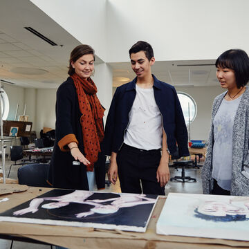 Three people in a large studio room looking at some canvases on a table. 