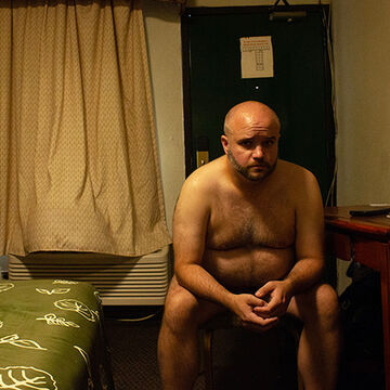 A naked person sits in a hotel room, looking at the camera