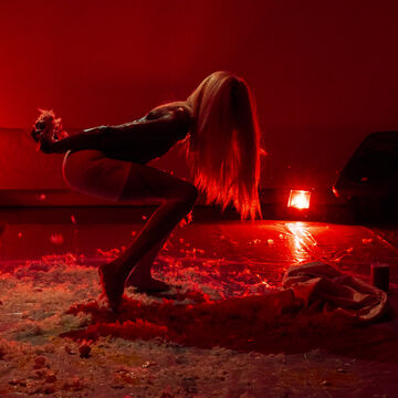 A person dances in a red-lit room with a floor of feathers