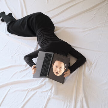 a person holding a laptop covering their head. On the screen is the person’s face.
