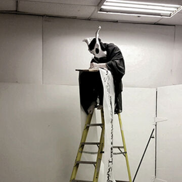 a person in an insectoid mask stands atop a ladder, writing on a piece of paper.