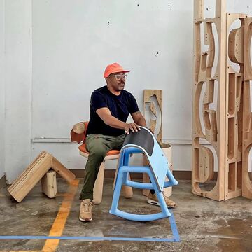 An image of Norman Teague sitting in his studio surrounded by large wooden pieces. 