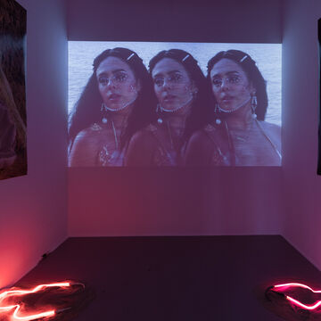 A dark room with photographs along the walls, pink neon on the floor and a video playing.