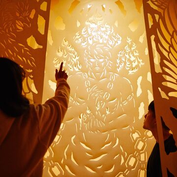 An image of two people observing an orange paper cut out piece of art with a soft white light.