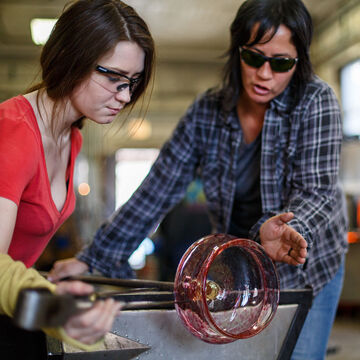 A faculty member assists a student in glassblowing