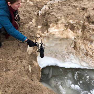 Katie Wood standing on a bank of sand holding a mic to a pool of frozen water.