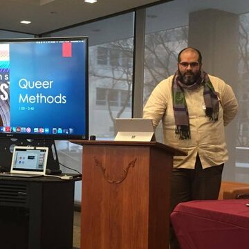 A person standing indoors at a podium. In the foreground, a computer monitor reads "Queer Methods" 