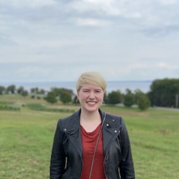 Susan Bay stands in a green field with a lake in the distance. She wears a cowl neck shirt and a leather jacket. Her hair is blond and cut short.