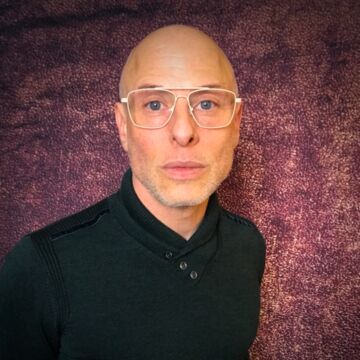 Joseph stands in front of a mottled purple backdrop, wearing a black shirt and glasses. They are bald.