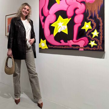 MJ stands beside a painting in a gallery. She has tight curls in her blonde hair and she wears a blazer over a white t-shirt.