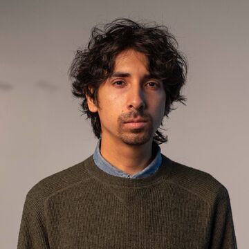 Juan is wearing a green ribbed sweater with the collar of a denim shirt peeking out at the neckline. He has a a mop of mussed up dark, wavy hair.