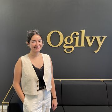 A woman stands wearing a cropped white vest and white pants poses in front of a sign that says "Ogilvy"