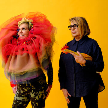 Two people standing against an orange background. The person on the right is holding a toy chicken.