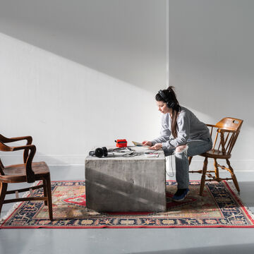 A person with headphones on sitting in a wooden chair and reading something over a block of cement on top of an area rug. An empty wooden chair and headphones sit across from them.