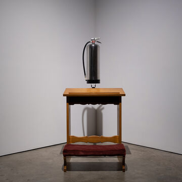 A sculpture of a chrome fire extinguisher suspended above a wooden piece of furniture, by Zach Dobbins