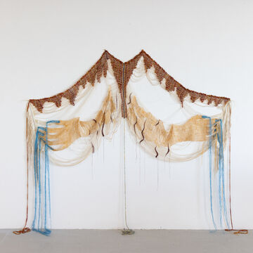 A wall hanging fiber art work of brown, beige and blue colors depicting two hands unfolding with blue nails represented with fringe.