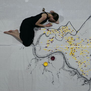 A photo of an adult in black clothing laying down nestled next to a minimally painted map with yellow and red finger marks, signification a specific location. The adult is looking down at the paint on their hand.