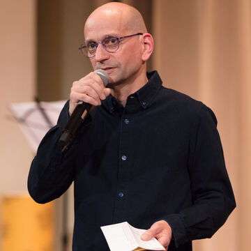 Still of an adult white man with glasses speaking into a microphone