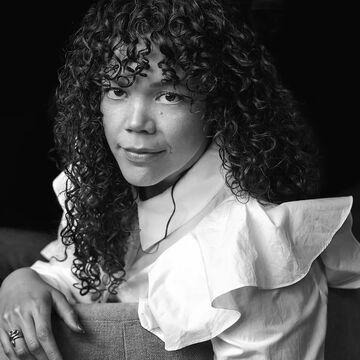A person with curly hair sits on a chair, wearing a white ruffled shirt, looking towards the camera. The image is in black and white.