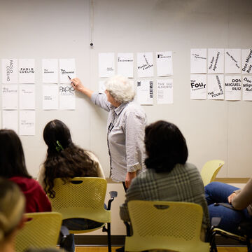An instructor stands in front of a classroom full of students pointing at a poster. 