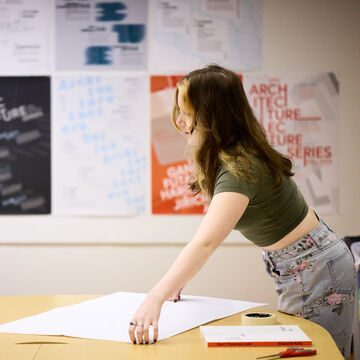 A student leans over a table in a classroom with a large piece of paper.