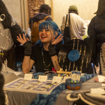 An Asian artist and white artist sit behind a table of jewelry and various artworks smiling and conversing with patrons.