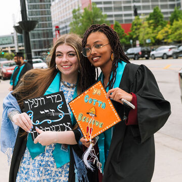 Two recent graduates—one with a fair skin tone and one with a medium-dark skin tone, smile and pose with their decorated graduation caps. 