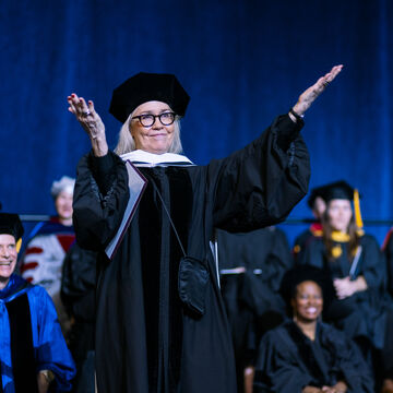 Anne Wilson, an adult white woman, wearing graduation garb and glasses, with raised open arms.