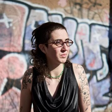 Portrait of Oona Taper, an adult person with a fair skintone, dark mullet, glasses and tattoos. 