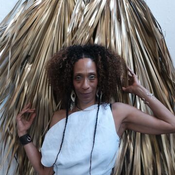 Portrait of Sonja Henderson, a Black woman with an afro and braids, posing in front of an artwork made of grass-like material.