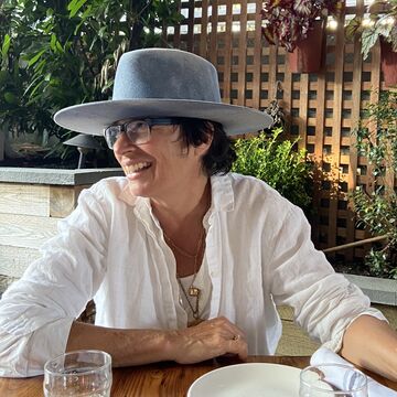 Portrait of Lora Fosberg, an adult person with a fair skin tone, short dark hair, glasses and a wide-brimmed hat.
