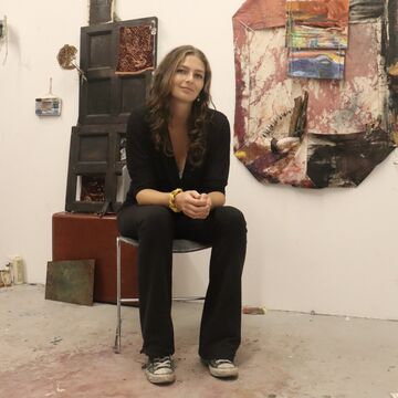 SAIC faculty member Charlotte Saylor, a person with light skin and brown hair, sits on a chair in front of artwork.