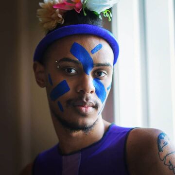 SAIC faculty member Compton Quashie, a Black artist wearing a blue shirt, and flowers in their hair.