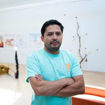 SAIC faculty member Juan Angel Chavez, a person with medium skin tone and dark hair, stands in front of a studio with artwork.