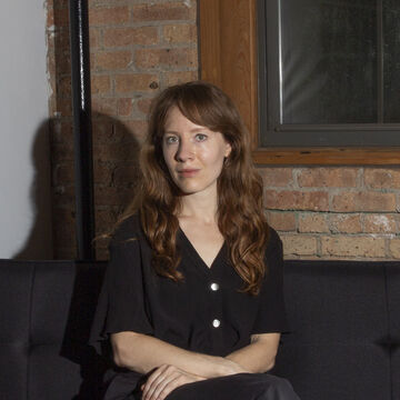 Kat Bawden, an adult person with a fair skin tone and wavy brown hair, sits on a couch indoors.