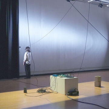 Mue standing in the performance studio (012),with their back facing the audience. There are multiple sound elements, including wires hanging from the ceiling and sound boards on pedestals. 