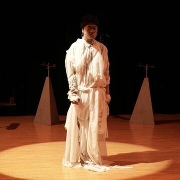 Performance artist Guanqiao surrounded in a circle of magnetic triangles, with a with tattered garment, and wire wrapped around their face. 
