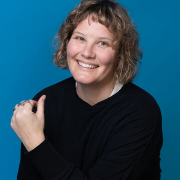 SAIC faculty member Sonja Thomsen, a person with light skin tone, and light curly hair, posing against a blue backdrop