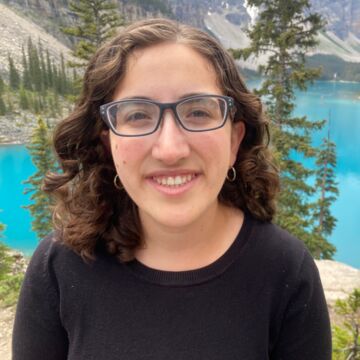 Headshot of Eva Shapiro, an adult person with fair skin, glasses, and curly shoulder-length brown hair.