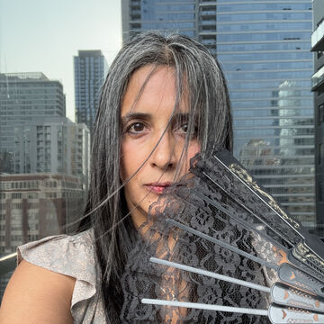 SAIC faculty member Gitanjali Kapila, a person with medium-light skin-tone and long, dark hair, stands outside with a fan.