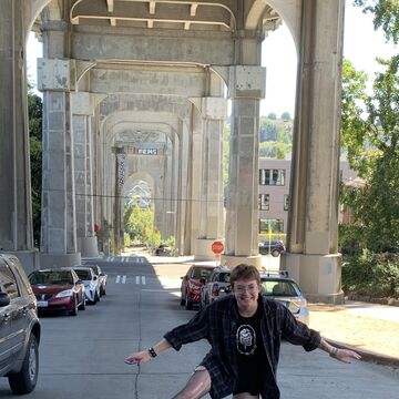 Portrait of Elliot O'Rourke, a white adult with short hair balancing on one foot in front of parked cars.