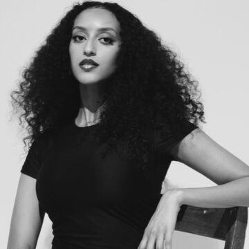 Black and white headshot of Billion Tekleab, an adult Black person with long curly hair and a medium-dark skin tone. 