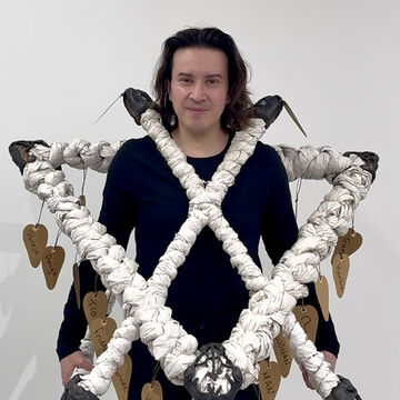 Portrait of Luisé Cisneros, an adult person with a medium-fair skin tone and straight brown hair, wearing a sculpture featuring tags with identifying phrases.