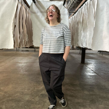Beth Sheehan, an adult person with a fair skin tone, shoulder-length blonde hair, and glasses, laughing and standing in a paper mill. 