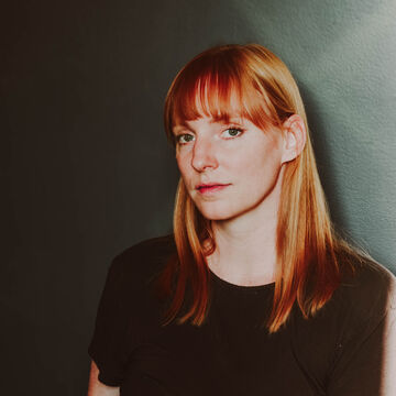 SAIC faculty member Kristin McWharter, a person with light skin tone and red hair, standing against a dark backdrop.