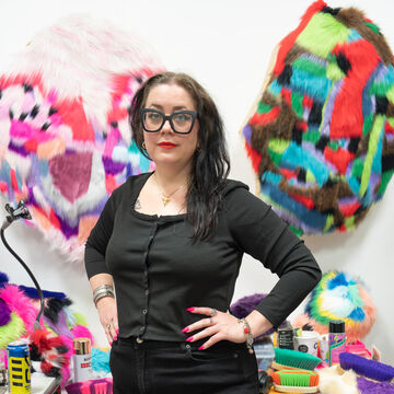 Portrait of Catie Burrill, an adult person with a fair skin tone, glasses, and dark hair, standing in a studio space. 