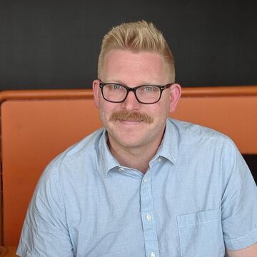 Headshot of Troy Briggs, an adult person with a fair skin tone, glasses, and short blonde hair. 