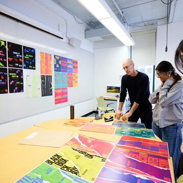 Two students and an instructor stand over a wooden table covered in bright graphics.