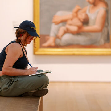 A person with light skin tone wearing a baseball hat sits on a bench with a sketchpad in front of a painting.